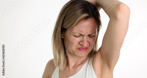 Young woman in white shirt stands on light background sniffing armpit. Lady winces at bad body odor. Bad smell of sweat. Chronic diseases or bad-quality deodorant