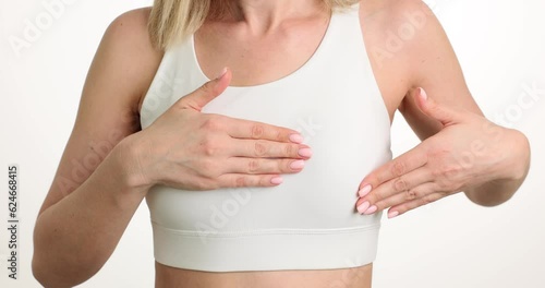 Female person in white top carefully touches chest to avoid pain standing on light background. Woman suffers from ache in breast. Breast cancer in women photo