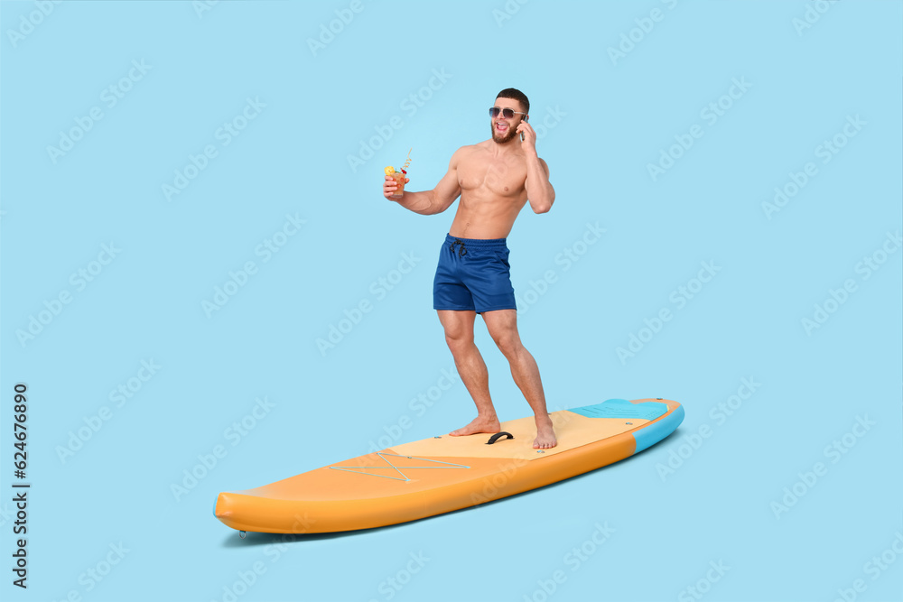 Happy man with refreshing drink talking on smartphone on SUP board against light blue background