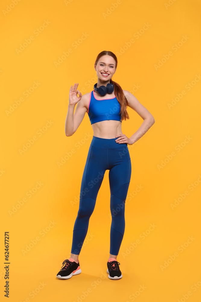 Young woman in sportswear and headphones showing ok gesture on yellow background Young woman in sportswear and headphones showing ok gesture on yellow background