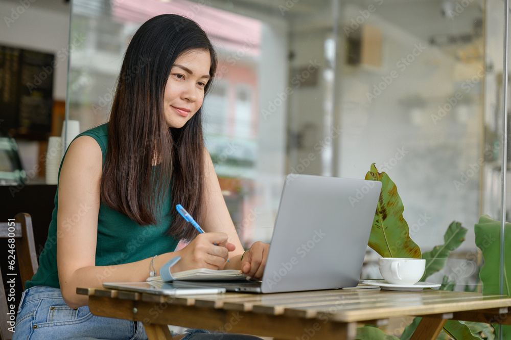 Portrait of Asian woman doing freelance work Young woman at cafe drinking coffee sitting at wooden table with laptop watching webinar and writing notes Portrait of Asian woman doing freelance work Young woman at cafe drinking coffee sitting at wooden table with laptop watching webinar and writing notes