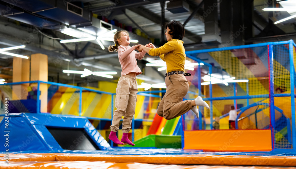 Pretty girl and her daughter kid jumping on colorful trampoline at playground park and smiling. Caucasian family happy during active entertaiments - obrazy, fototapety, plakaty 