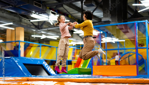 Pretty girl and her daughter kid jumping on colorful trampoline at playground park and smiling. Caucasian family happy during active entertaiments photo
