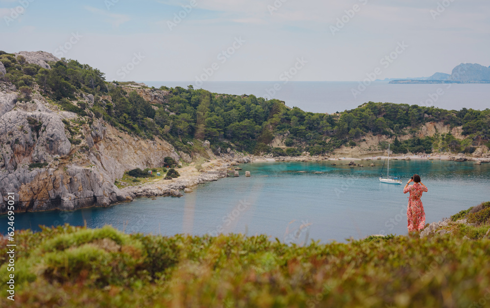 Fototapeta premium Asian woman in hat look on views of azure Bay in Mediterranean sea. Travel and vacation concept. Anthony Quinn bay with crystal clear water in Rhodes island, Greece. The most beautiful beach.