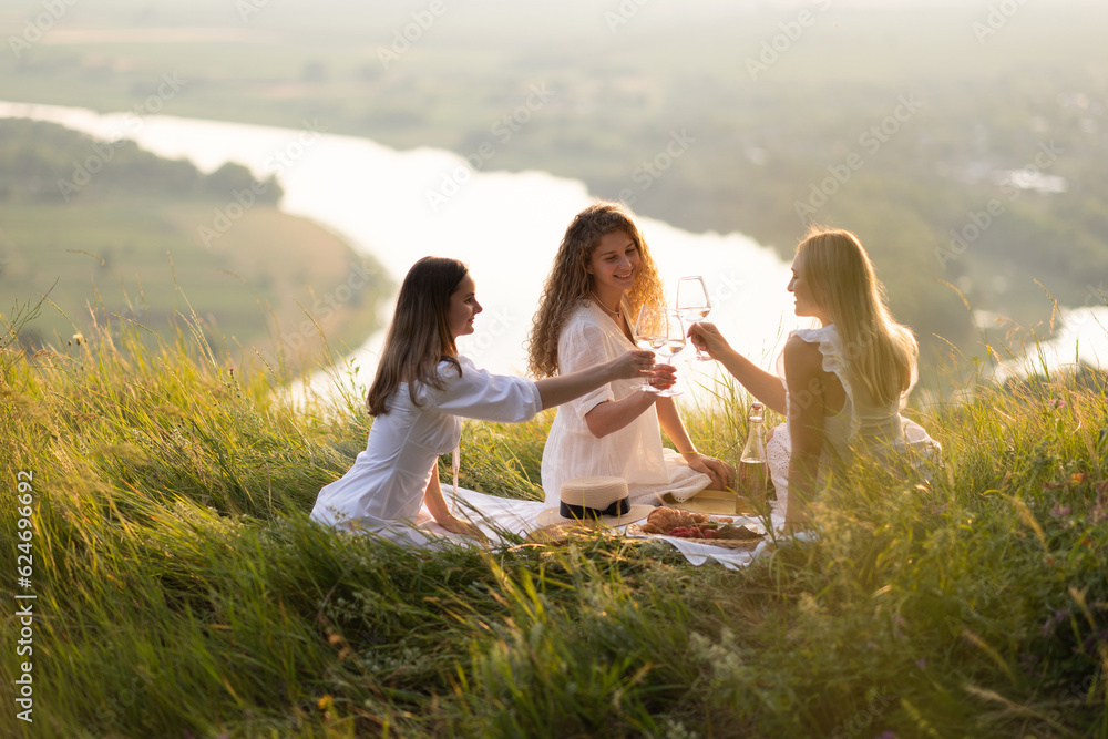 Beautiful girlfriends having picnic on the hill on summer sunset. They sitting with hands up and enjoying beautiful landscape.