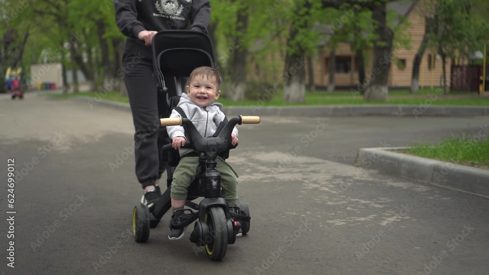 Obraz premium A young mother carries a small child on a tricycle. Two-year-old boy on a bicycle close-up.
