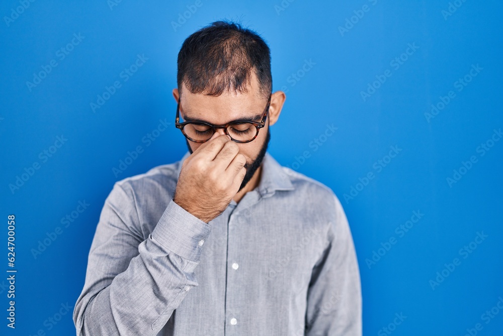 Middle east man with beard standing over blue background tired rubbing nose and eyes feeling fatigue and headache. stress and frustration concept. Middle east man with beard standing over blue background tired rubbing nose and eyes feeling fatigue and headache. stress and frustration concept.