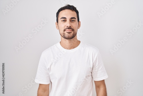 Handsome hispanic man standing over white background relaxed with serious expression on face. simple and natural looking at the camera.