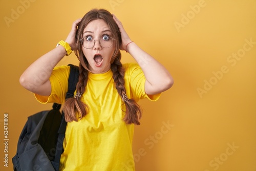 Young caucasian woman wearing student backpack over yellow background crazy and scared with hands on head, afraid and surprised of shock with open mouth