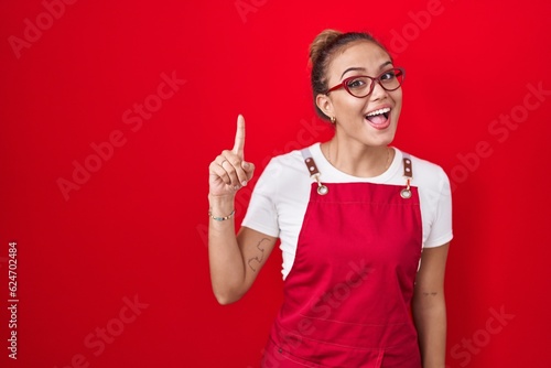 Young hispanic woman wearing waitress apron over red background pointing finger up with successful idea. exited and happy. number one. photo
