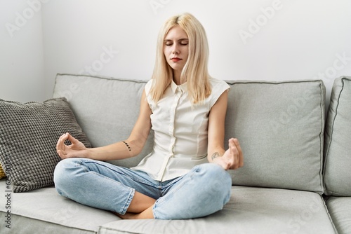 Young blonde woman doing yoga exercise sitting on sofa at home