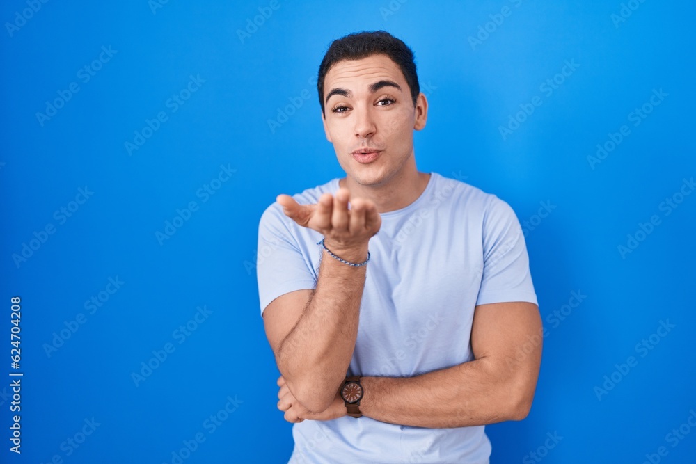 Young hispanic man standing over blue background looking at the camera blowing a kiss with hand on air being lovely and sexy. love expression. Young hispanic man standing over blue background looking at the camera blowing a kiss with hand on air being lovely and sexy. love expression.