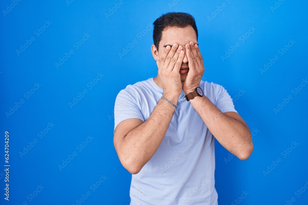 Young hispanic man standing over blue background rubbing eyes for fatigue and headache, sleepy and tired expression. vision problem Young hispanic man standing over blue background rubbing eyes for fatigue and headache, sleepy and tired expression. vision problem