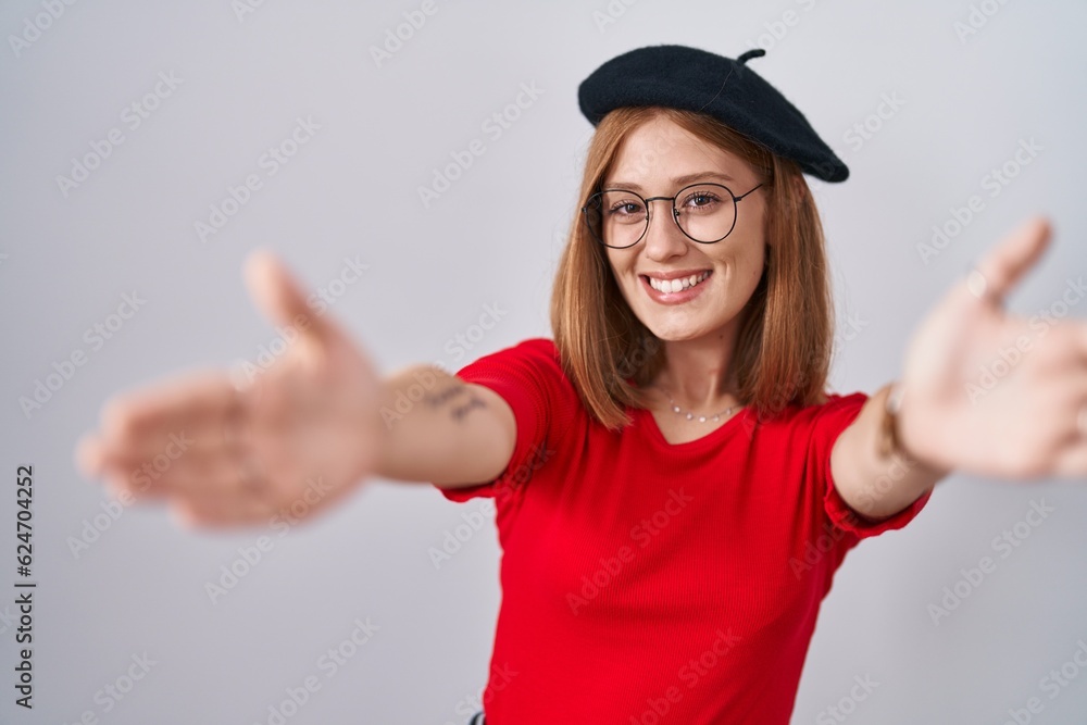 Young redhead woman standing wearing glasses and beret looking at the camera smiling with open arms for hug. cheerful expression embracing happiness. Young redhead woman standing wearing glasses and beret looking at the camera smiling with open arms for hug. cheerful expression embracing happiness.
