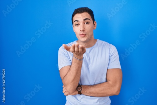 Young hispanic man standing over blue background looking at the camera blowing a kiss with hand on air being lovely and sexy. love expression.
