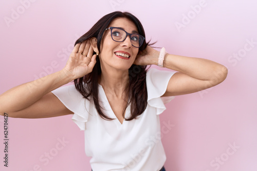 Middle age hispanic woman wearing casual white t shirt and glasses trying to hear both hands on ear gesture, curious for gossip. hearing problem, deaf