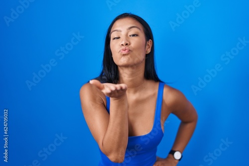 Hispanic woman standing over blue background looking at the camera blowing a kiss with hand on air being lovely and sexy. love expression.