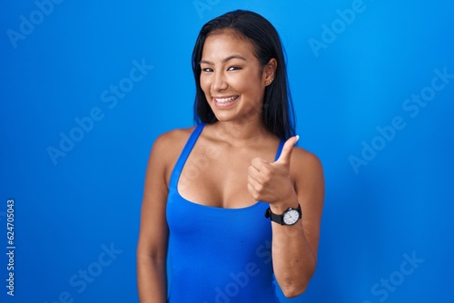 Hispanic woman standing over blue background doing happy thumbs up gesture with hand. approving expression looking at the camera showing success.