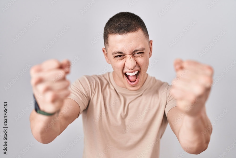 Young man standing over isolated background angry and mad raising fists frustrated and furious while shouting with anger. rage and aggressive concept. Young man standing over isolated background angry and mad raising fists frustrated and furious while shouting with anger. rage and aggressive concept.