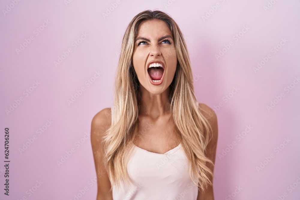 Young blonde woman standing over pink background angry and mad screaming frustrated and furious, shouting with anger. rage and aggressive concept. Young blonde woman standing over pink background angry and mad screaming frustrated and furious, shouting with anger. rage and aggressive concept.