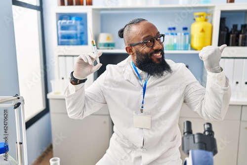 African american man working at scientist laboratory holding syringe pointing thumb up to the side smiling happy with open mouth