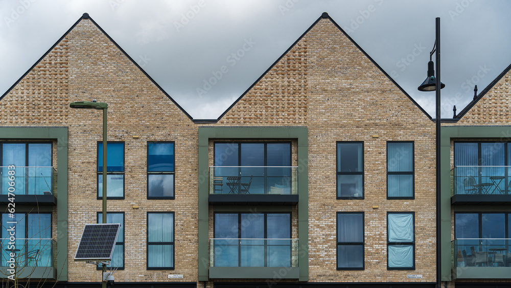 Symmetrical geometric rhythms on brick wall, triangular roofs, rectangular windows and balconies. Folding furniture on balconies. Two street lamps, solar panel in foreground. Cloudy English sky. Symmetrical geometric rhythms on brick wall, triangular roofs, rectangular windows and balconies. Folding furniture on balconies. Two street lamps, solar panel in foreground. Cloudy English sky.