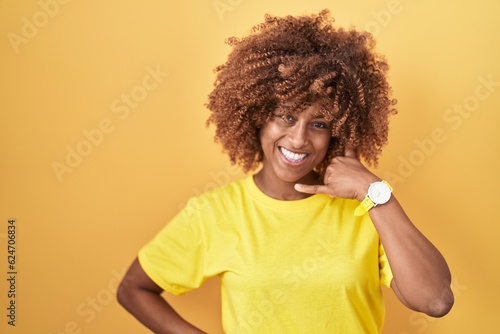 Young hispanic woman with curly hair standing over yellow background smiling doing phone gesture with hand and fingers like talking on the telephone. communicating concepts.
