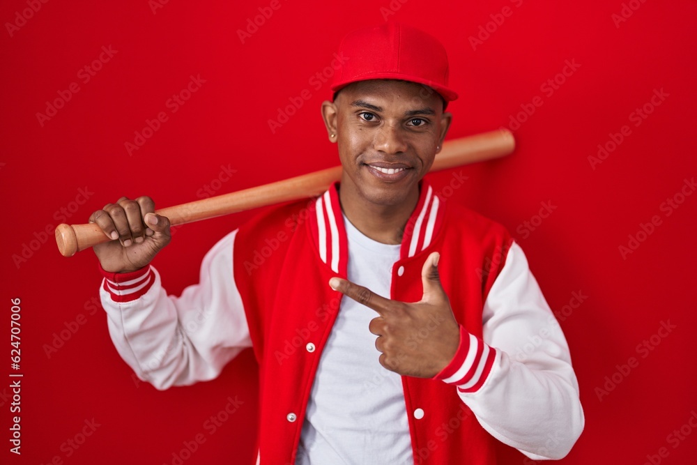 Young hispanic man playing baseball holding bat cheerful with a smile on face pointing with hand and finger up to the side with happy and natural expression Young hispanic man playing baseball holding bat cheerful with a smile on face pointing with hand and finger up to the side with happy and natural expression