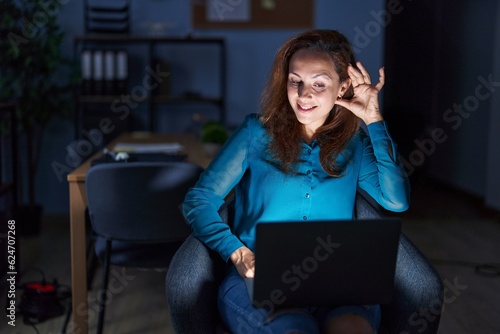 Brunette woman working at the office at night smiling with hand over ear listening an hearing to rumor or gossip. deafness concept.