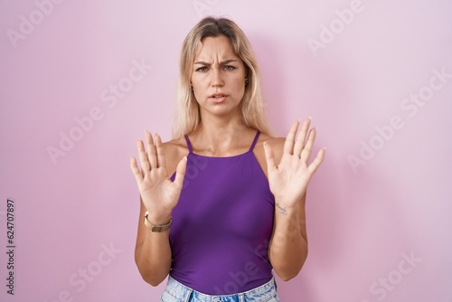Young blonde woman standing over pink background moving away hands palms showing refusal and denial with afraid and disgusting expression. stop and forbidden.