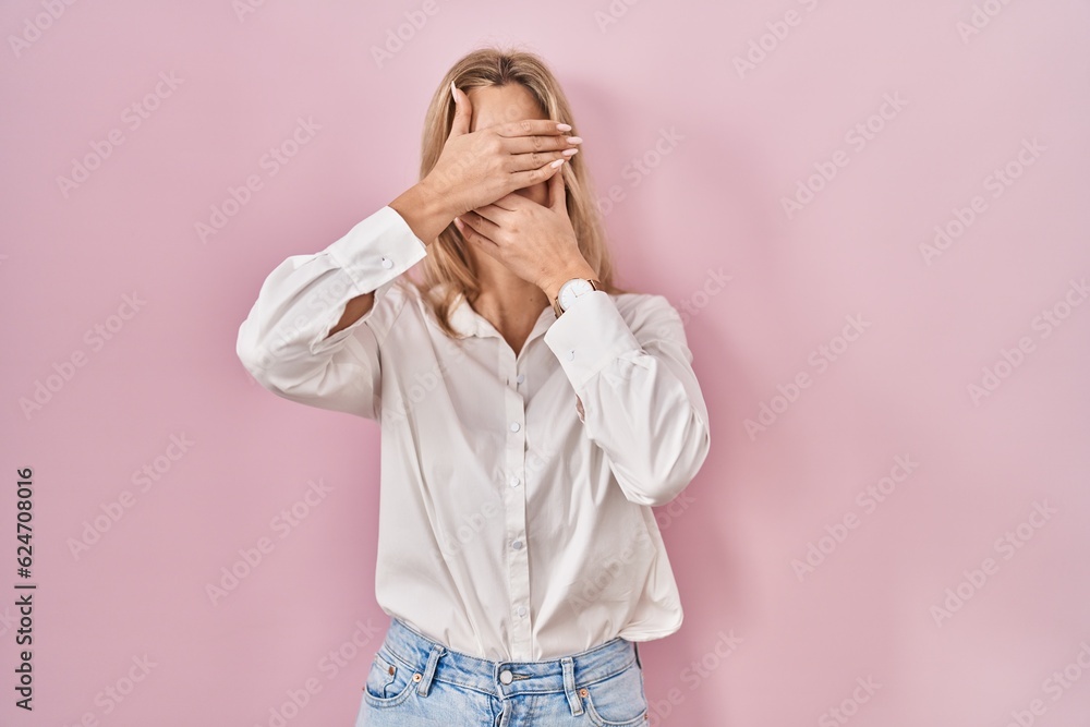 Young caucasian woman wearing casual white shirt over pink background covering eyes and mouth with hands, surprised and shocked. hiding emotion Young caucasian woman wearing casual white shirt over pink background covering eyes and mouth with hands, surprised and shocked. hiding emotion