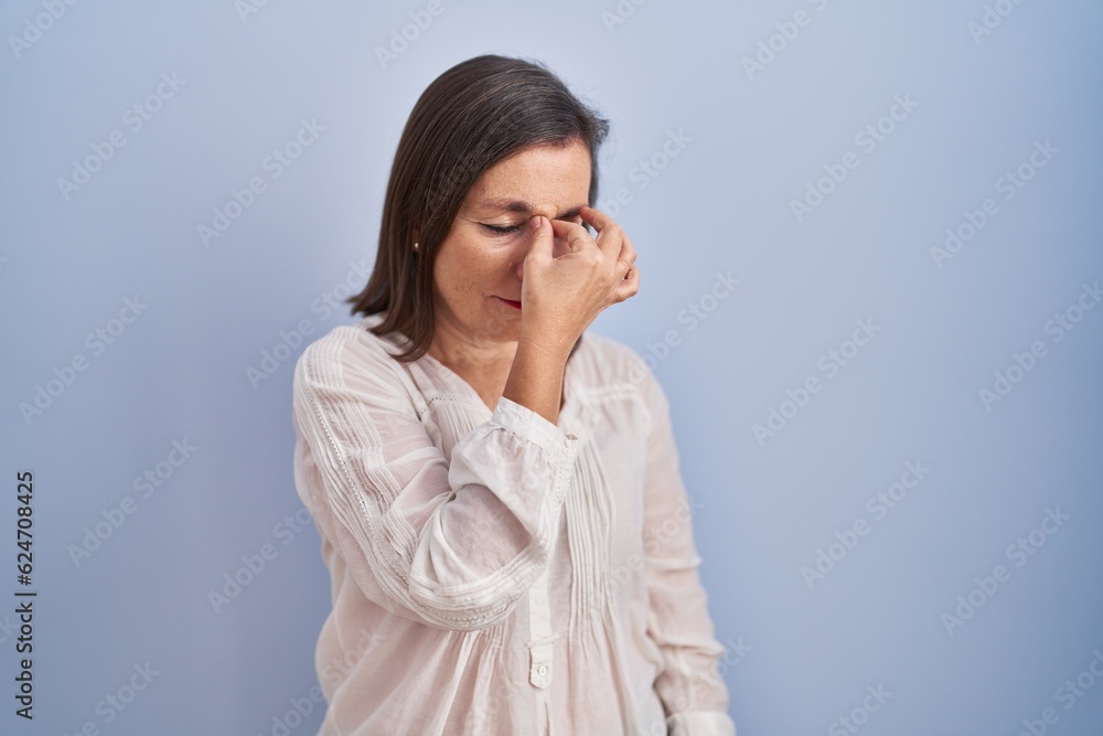 Middle age hispanic woman standing over blue background tired rubbing nose and eyes feeling fatigue and headache. stress and frustration concept. Middle age hispanic woman standing over blue background tired rubbing nose and eyes feeling fatigue and headache. stress and frustration concept.