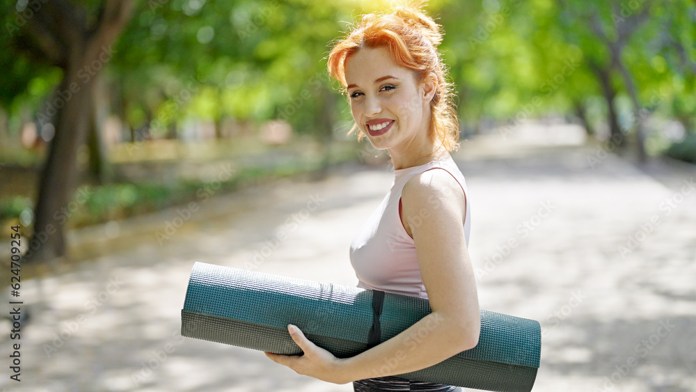 Naklejka premium Young redhead woman smiling wearing sportswear holding yoga mat at park