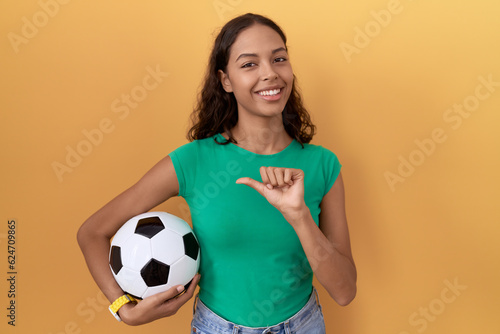 Young hispanic woman holding ball pointing to the back behind with hand and thumbs up  smiling confident
