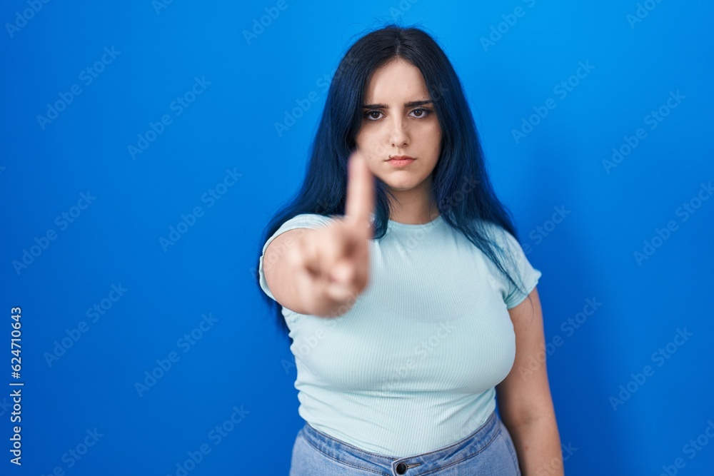 Fototapeta premium Young modern girl with blue hair standing over blue background pointing with finger up and angry expression, showing no gesture