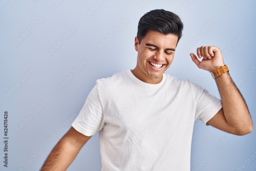 Hispanic man standing over blue background dancing happy and cheerful, smiling moving casual and confident listening to music Hispanic man standing over blue background dancing happy and cheerful, smiling moving casual and confident listening to music