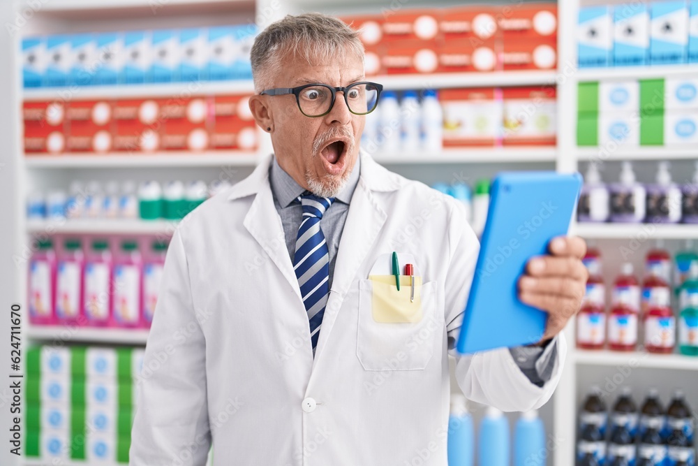 Hispanic man with grey hair working at pharmacy drugstore doing video call with tablet scared and amazed with open mouth for surprise, disbelief face