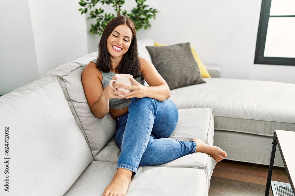Young beautiful hispanic woman drinking coffee sitting on sofa at home Young beautiful hispanic woman drinking coffee sitting on sofa at home
