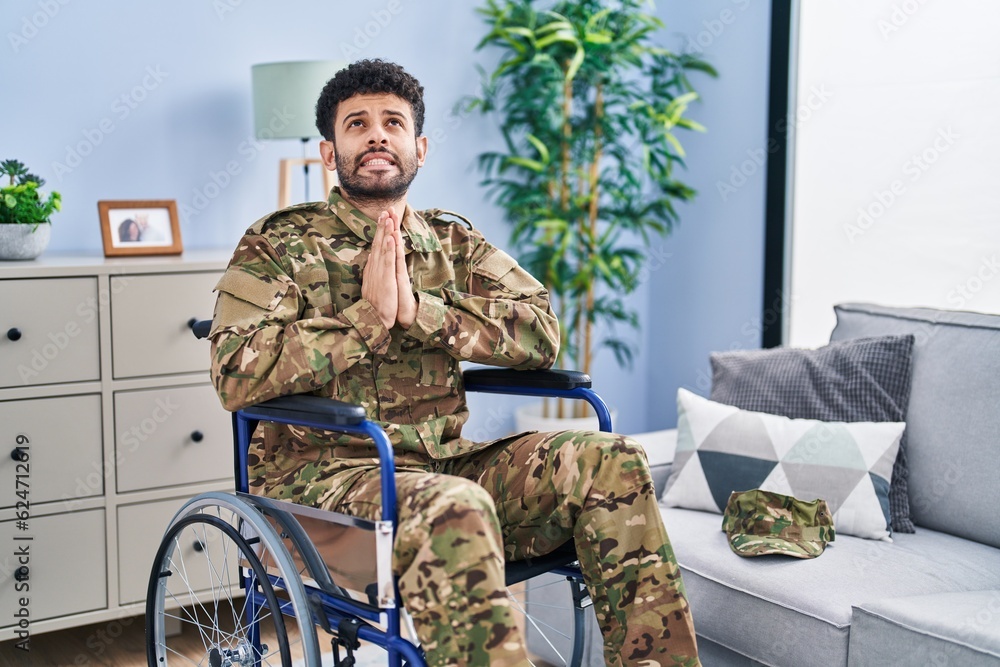 Arab man wearing camouflage army uniform sitting on wheelchair begging and praying with hands together with hope expression on face very emotional and worried. begging.