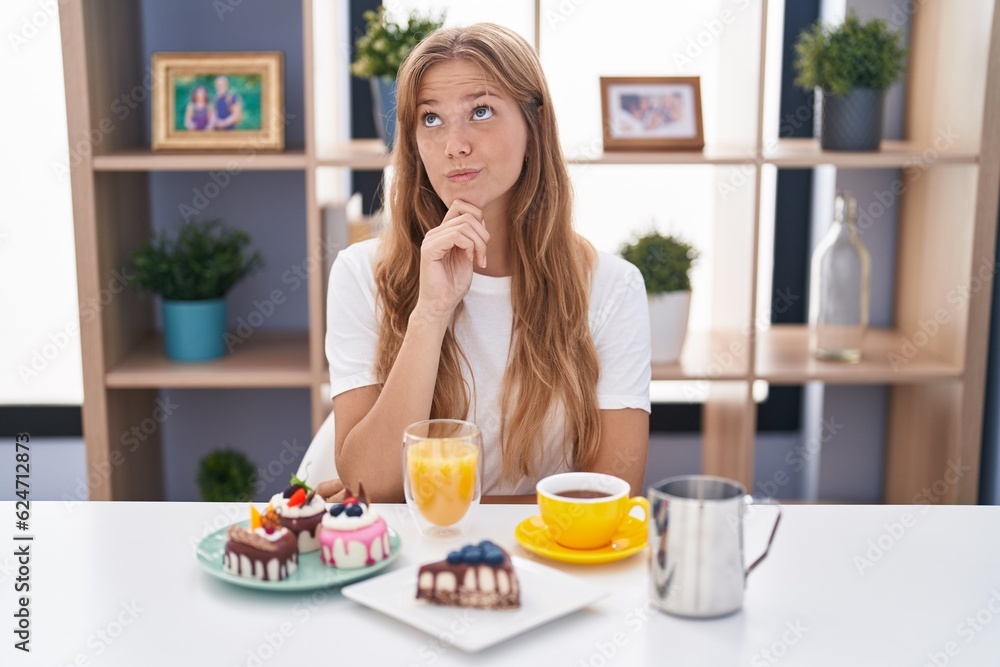Young caucasian woman eating pastries t for breakfast with hand on chin thinking about question, pensive expression. smiling with thoughtful face. doubt concept. Young caucasian woman eating pastries t for breakfast with hand on chin thinking about question, pensive expression. smiling with thoughtful face. doubt concept.