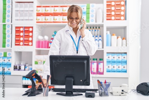 Young blonde girl pharmacist talking on smartphone using computer at pharmacy