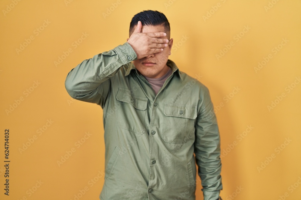 Hispanic young man standing over yellow background covering eyes with hand, looking serious and sad. sightless, hiding and rejection concept Hispanic young man standing over yellow background covering eyes with hand, looking serious and sad. sightless, hiding and rejection concept
