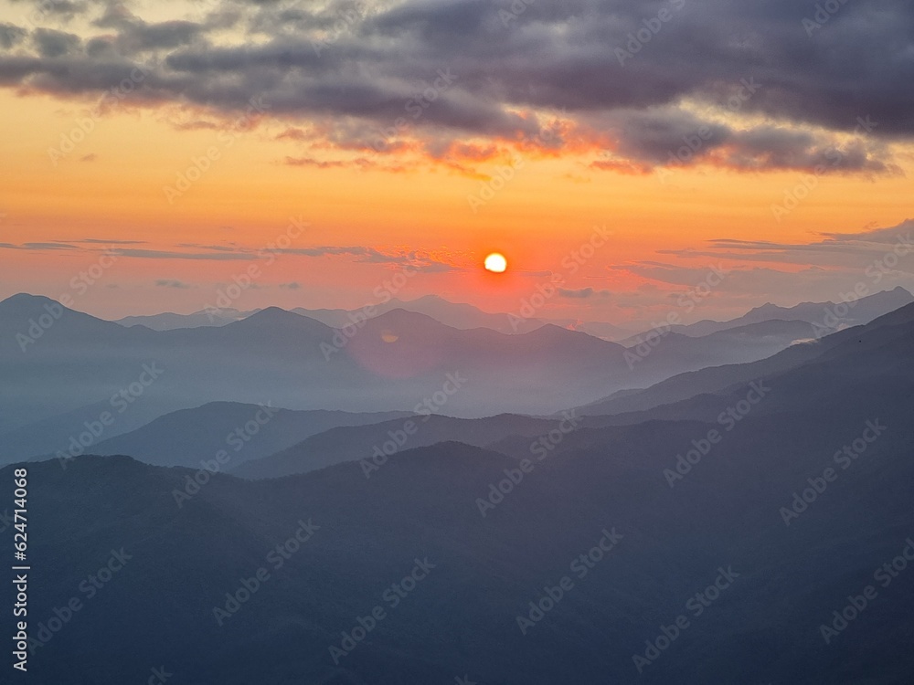 
The sunset in the mountains unveils a spectacular panorama, as the sun's crimson farewell stains the sky, casting ethereal shadows over the rugged peaks.