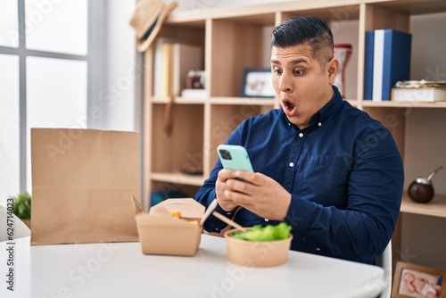 Hispanic young man eating take away food using smartphone afraid and shocked with surprise and amazed expression, fear and excited face.