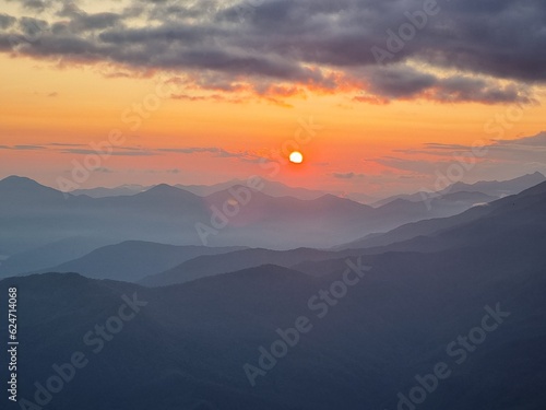  The sunset in the mountains unveils a spectacular panorama, as the sun's crimson farewell stains the sky, casting ethereal shadows over the rugged peaks.