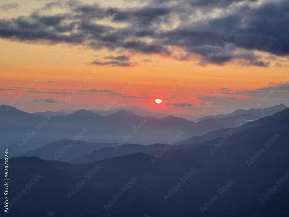 Fototapeta premium The sun dips below the mountain horizon, its fiery descent casting a rich palette of golds and purples across the sky, embodying the tranquil majesty of a mountain sunset.
