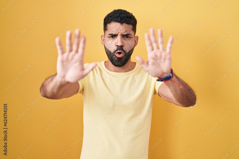 Fototapeta premium Hispanic man with beard standing over yellow background doing stop gesture with hands palms, angry and frustration expression