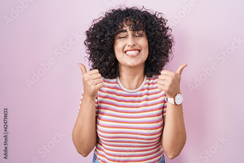 Young middle east woman standing over pink background success sign doing positive gesture with hand, thumbs up smiling and happy. cheerful expression and winner gesture.