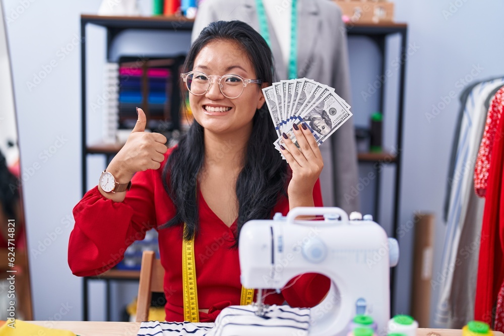 Asian young woman dressmaker designer holding dollars smiling happy and positive, thumb up doing excellent and approval sign