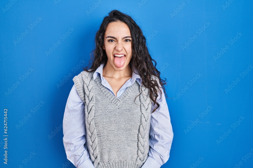 Young brunette woman standing over blue background sticking tongue out happy with funny expression. emotion concept. Young brunette woman standing over blue background sticking tongue out happy with funny expression. emotion concept.